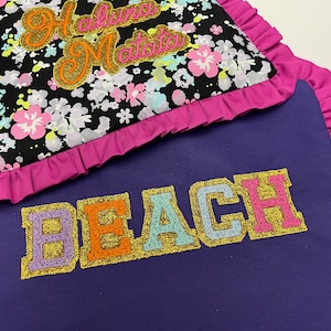 May include: A purple fabric pillow with pink ruffled edges and the word "BEACH" embroidered in colorful glittery letters. A smaller pillow with a black background and pink ruffled edges is on top of the larger pillow. The smaller pillow has the words "Hakuna Matata" embroidered in gold glitter.