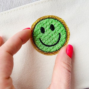 May include: A green smiley face embroidered patch with a gold outline, sewn onto a white canvas fabric.