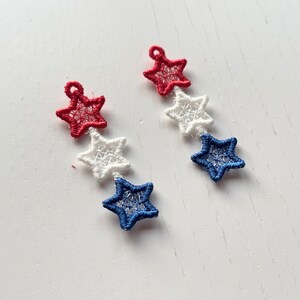 Pu&ograve; includere: Un paio di orecchini con tre stelle ciascuno. Le stelle sono rosse, bianche e blu.