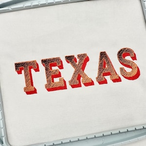 May include: Embroidered design of the word "TEXAS" in a bold, block letter style. The letters are a gradient of red and orange, with a textured appearance. The design is set against a white background and framed by a light blue embroidery hoop.