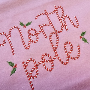 Puede incluir: Camiseta rosa con "Polo Norte" bordado en rayas de bastón de caramelo rojo y blanco. Hojas de acebo verdes están bordadas a cada lado del texto.
