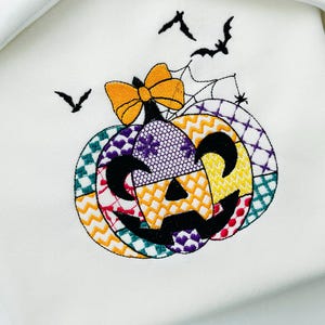 May include: Embroidered Halloween design featuring a patchwork pumpkin with a black face, an orange bow, and a spiderweb. Three black bats fly above the pumpkin on a white background. The design is likely for a seasonal craft or apparel item.