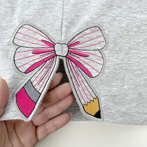 Puede incluir: Una tela jaspeada de color gris claro con una aplicación bordada en forma de lazo. El lazo está formado por dos lápices con gomas de borrar rosas, virolas grises y puntas amarillas, acentuado con finas líneas rosas y un nudo central blanco.