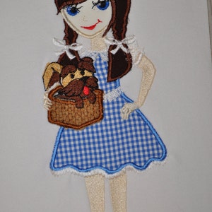 May include: Embroidered patch of a girl in a blue and white gingham dress holding a brown dog in a basket. She has brown hair, blue eyes, and red shoes.