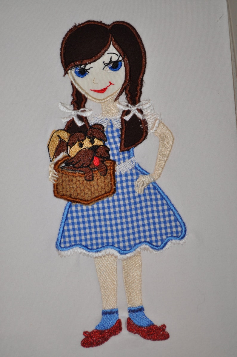 May include: Embroidered patch of a girl in a blue and white gingham dress holding a brown dog in a basket. She has brown hair, blue eyes, and red shoes.