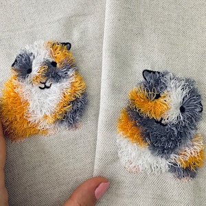 May include: Two embroidered guinea pigs on a beige fabric background. One guinea pig is orange, white, and black. The other guinea pig is yellow, white, and gray.