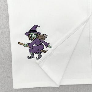 May include: A white kitchen towel featuring an embroidered graphic of a green-skinned witch with brown hair, wearing a purple dress and a matching pointed hat, riding a brown broomstick. The fabric has a visible woven texture throughout the cloth.