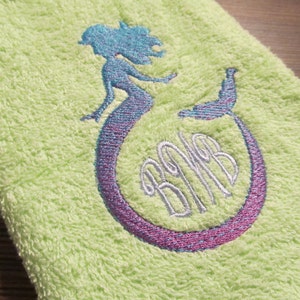 以下が含まれることがあります： 薄い緑色のタオルに、紫と青の刺繍で人魚のデザインが施されています。人魚の尻尾は円形に巻かれ、銀色の糸で「BMB」のモノグラムが刺繍されています。