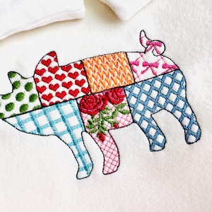 Op de afbeelding: Een patchwork varkensontwerp geborduurd op een witte stof. Het varken is gemaakt van verschillende gekleurde vierkanten met patronen zoals harten, bloemen en ruiten.