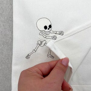 May include: A white kitchen towel featuring a black embroidered skeleton graphic. The skeleton is depicted in a dynamic pose with arms extended forward. The fabric has a visible herringbone weave pattern throughout the surface of the textile.