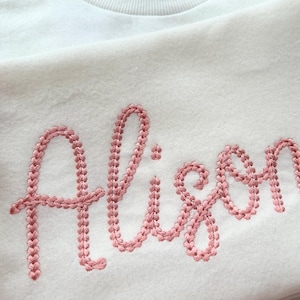 Puede incluir: Una sudadera blanca con el nombre "Alison" bordado en rosa.