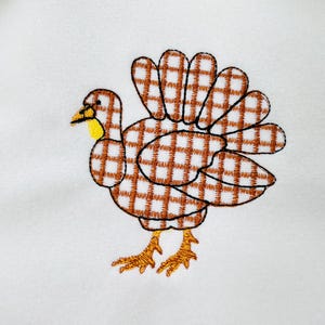 May include: Embroidered turkey design with brown and white plaid pattern on a white background. The turkey has a yellow beak and feet. The design is likely for a Thanksgiving or fall-themed product.