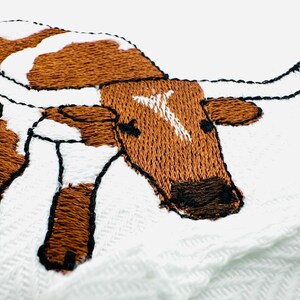 May include: Embroidered image of a longhorn steer with brown and white markings. The steer has large curved horns and is stitched with brown and black thread on a white background. The embroidery is detailed and textured.