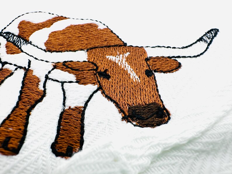 May include: Embroidered image of a longhorn steer with brown and white markings. The steer has large curved horns and is stitched with brown and black thread on a white background. The embroidery is detailed and textured.