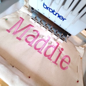 May include: A close-up of a white embroidery hoop with the name "Maddie" embroidered in pink thread. The embroidery machine is in the background.
