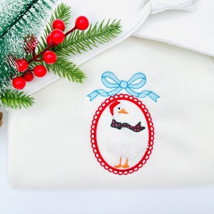 May include: White blanket with an embroidered Christmas goose wearing a Santa hat and scarf, framed in a red oval with a blue bow. A sprig of evergreen with red berries and a pinecone are in the upper left corner.