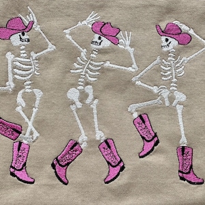 May include: Embroidered design featuring three dancing skeletons wearing pink cowboy hats and boots. The skeletons are white, with pink accents on the hats and boots, set against a tan background.