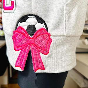Könnte beinhalten: Ein grauer Hoodie mit einem pinkfarbenen und weißen Fußball und einer pinkfarbenen Schleife am Ärmel.