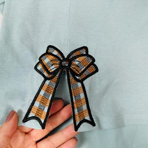 May include: A light blue shirt with a brown and black plaid bow applique.