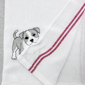 May include: White kitchen towel with an embroidered image of a gray and white puppy. The towel has two vertical red stripes on the edge.