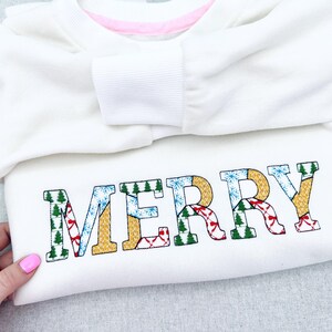 Könnte beinhalten: Weißes Sweatshirt mit dem Wort "MERRY", das mit farbenfrohen Urlaubsmustern bestickt ist. Die Buchstaben zeigen Weihnachtsbäume, Schneeflocken und rote Schleifen. Das Sweatshirt hat einen rosa Ausschnitt.