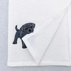 May include: White waffle-weave towel with an embroidered black puppy. The puppy is depicted in a standing pose. The towel has a folded corner, showing its texture. The background is a plain light grey.
