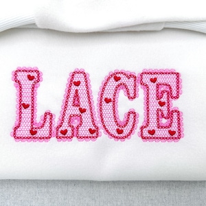 Op de afbeelding: Witte stof met het woord "LACE" geborduurd in roze. De letters zijn omlijnd met een geschulpte rand en gevuld met een kantpatroon. Kleine rode hartjes zijn verspreid in elke letter.