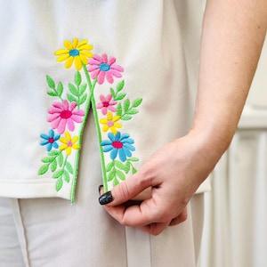 May include: White garment with a colourful floral embroidery design. The embroidery features yellow, pink, and blue flowers with green leaves. The garment is being held.