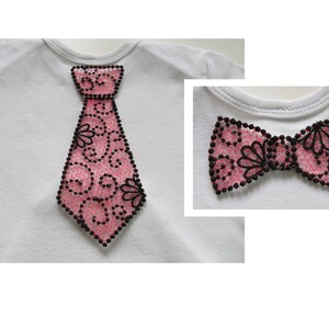 Puede incluir: Una camiseta blanca con una aplicación de corbata rosa y negra. La corbata tiene un diseño decorativo de espirales y flores. Una imagen separada muestra una pajarita rosa y negra con el mismo diseño.