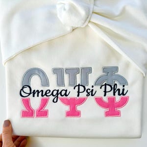 May include: White hooded sweatshirt with the Greek letters Omega Psi Phi embroidered in gray and pink.