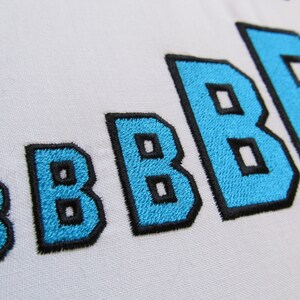 Puede incluir: Primer plano de cinco letras bordadas que deletrean "BBBB" en azul con contornos negros. Las letras son de diferentes tamaños, siendo la más grande a la derecha y la más pequeña a la izquierda.
