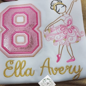 May include: A white shirt with pink and gold embroidery of a ballerina, the number 8, and the names "Ella Avery".