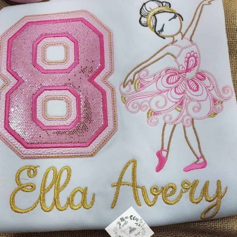 May include: A white shirt with pink and gold embroidery of a ballerina, the number 8, and the names "Ella Avery".