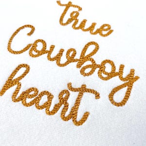 May include: White fabric with gold embroidered text that reads "True Cowboy Heart" in a cursive font. The embroidery has a textured appearance, and the letters are slightly spaced.
