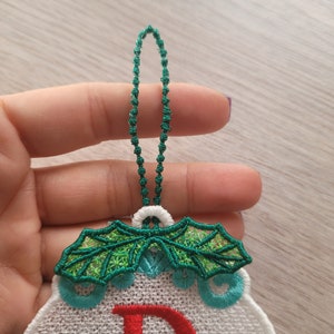 May include: White fabric Christmas ornament with green holly leaves and a red letter D. The ornament has a green beaded loop for hanging.