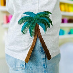以下が含まれることがあります： 白地に茶色と緑の刺繍でヤシの木がデザインされたTシャツ。青いジーンズの上に着ています。