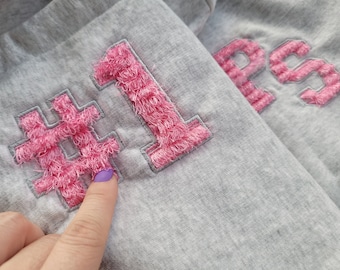 May include: Grey sweatshirt with pink fuzzy embroidered text that reads "#1s".