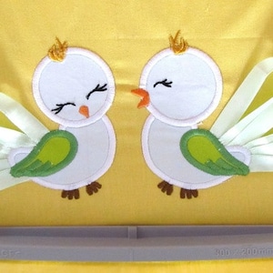 May include: Two embroidered bird appliqués on a yellow background. The birds have white bodies, green wings, and ribbon tail feathers. Each bird wears a gold crown. The birds are facing each other.