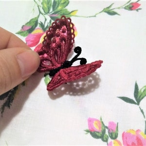 以下が含まれることがあります： 黒色の触角を持つ、ワインレッドのクロシェ編み蝶。蝶は、花柄の白い背景の上にあります。