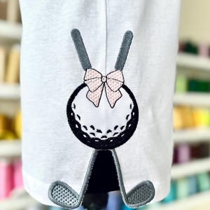 May include: A white golf ball with a black outline and a pink bow on top. The golf ball is sitting on top of two black golf clubs.