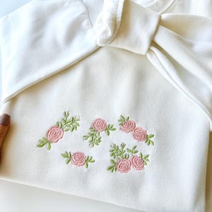 Puede incluir: Sudadera blanca con rosas bordadas de color rosa y hojas verdes.