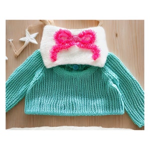 Puede incluir: Un suéter de punto turquesa con cuello blanco y un lazo rosa.