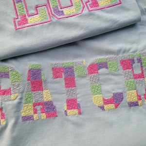 May include: A light blue t-shirt with the word "PATCH" embroidered in a checkered pattern using pink, yellow, green, purple, and gray thread.