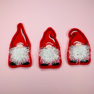 May include: Three red felt gnome ornaments with white fuzzy beards. The gnomes are standing upright with their arms at their sides.