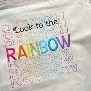 May include: A white tote bag with the text "Look to the rainbow" embroidered in rainbow colors. The text is repeated multiple times in a stacked pattern, with each repetition slightly smaller than the one above it.