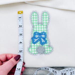 Puede incluir: Aplicación bordada de un conejito sobre tela blanca. El conejito es de cuadros verdes y blancos con un lazo azul y detalles plateados. El conejito mide unos 15 cm de alto.