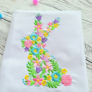 May include: White fabric with an embroidered floral design in the shape of a bunny. The flowers are pink, yellow, blue, green, and purple.