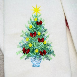 Op de afbeelding: Geborduurde kerstboomontwerp op een witte linnen theedoek. De boom is groen met gele ornamenten en rode strikken. De boom staat in een blauwe pot met een gele ster bovenop.