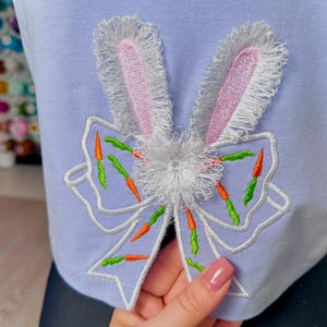 Può includere: T-shirt azzurra con un design ricamato di orecchie di coniglio e fiocco. Le orecchie di coniglio sono grigie con l'interno rosa e un bordo sfrangiato. Il fiocco è azzurro con accenti di carote arancioni e verdi e un centro bianco soffice.