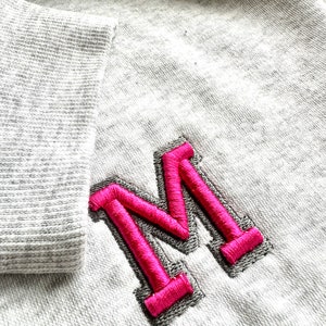 May include: A gray sweatshirt with a pink embroidered letter "M" on the front.